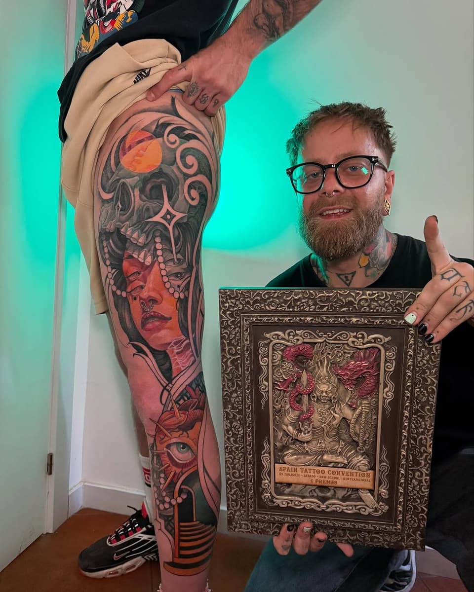 Tattoo artist displays full leg sleeve featuring a woman's face with skull imagery in red and black ink, holding a framed award.