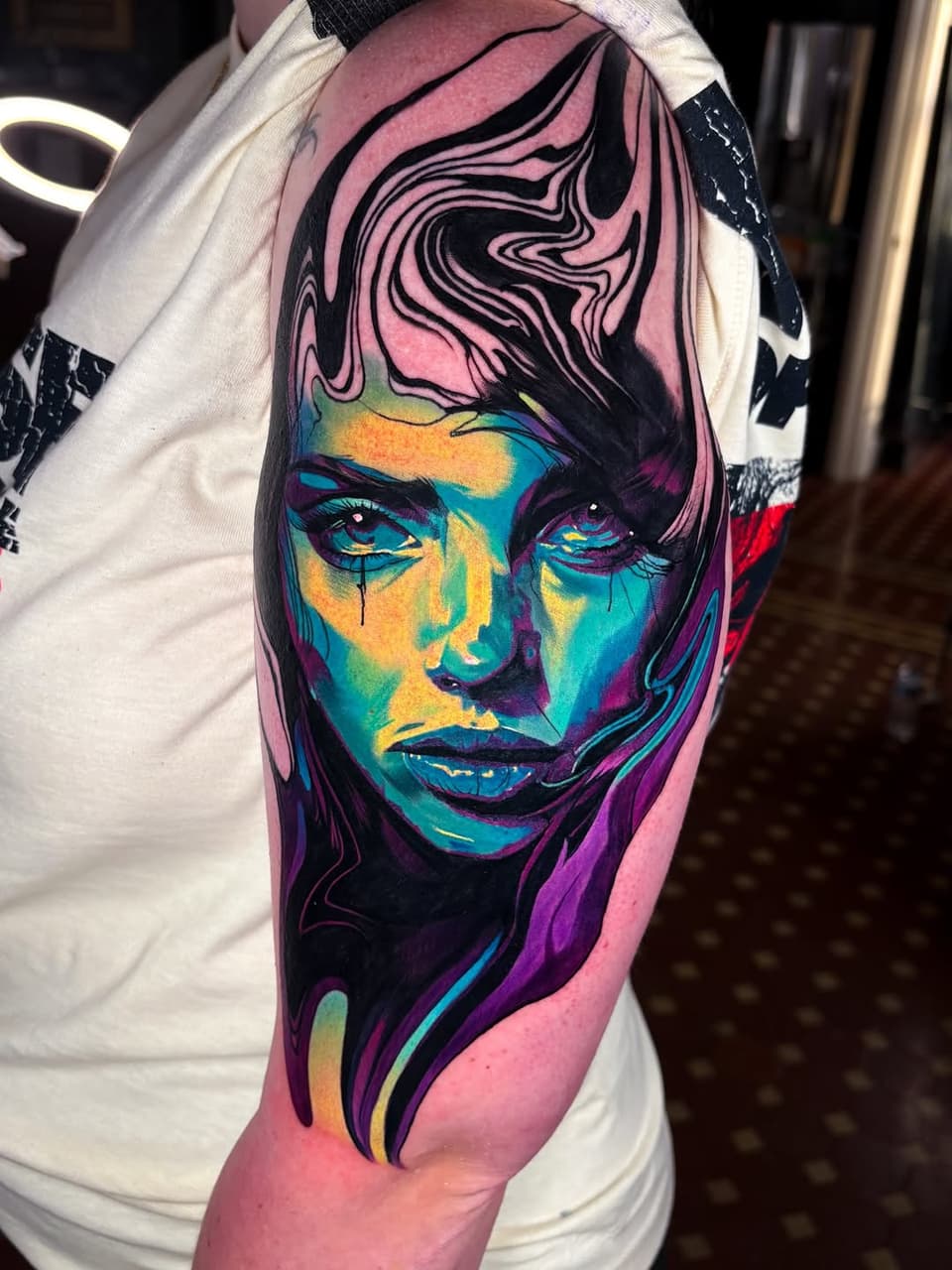 Colorful portrait tattoo of a person's face with vibrant blue, yellow, purple, and pink hues, located on the upper arm.