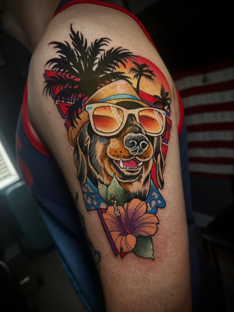 Colorful shoulder tattoo of a smiling dog wearing sunglasses and a tropical shirt with palm trees, flowers, and a sunset backdrop.