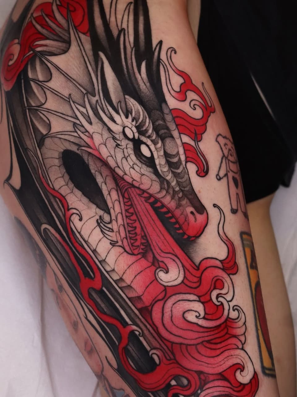 Dragon head tattoo on the leg featuring gray scales, red ornamental flames, and black line work with detailed shading and decorative scrollwork elements.
