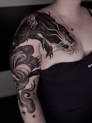 Dragon and female face tattoo on upper arm and shoulder in black ink with flowing hair details and stars.