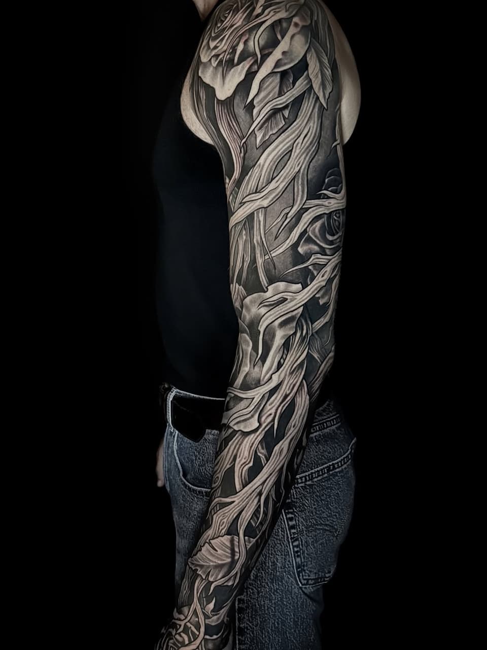 Full-length arm sleeve tattoo featuring flowing, layered organic forms in black and grey ink on a person wearing jeans.