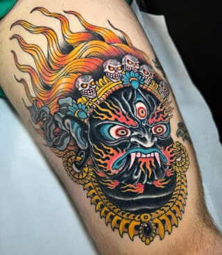 Colorful demon mask tattoo on upper leg featuring skulls, flames, multiple eyes, ornate gold collar, and blue floral accents.