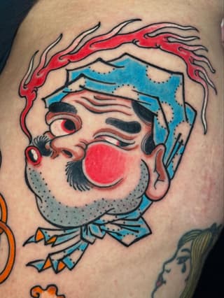Colorful clown head tattoo on arm featuring red nose, blue hat, red flames, and gray beard with black outlines and detailed facial features.