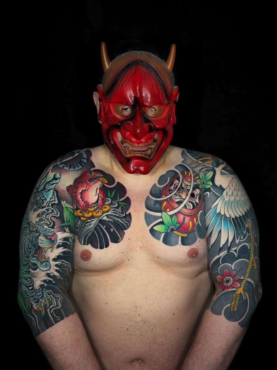 Shirtless person wearing full-color sleeve tattoos featuring demons, birds, and floral motifs in red, black, and green, paired with a red horned devil mask.