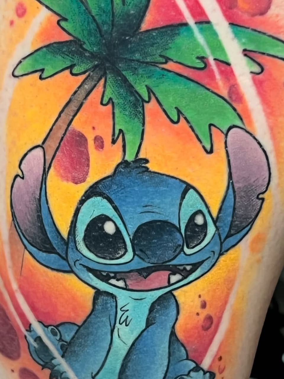 Colorful shoulder tattoo featuring a blue alien character with large eyes holding a green palm tree, surrounded by vibrant orange, yellow, red, and pink background.