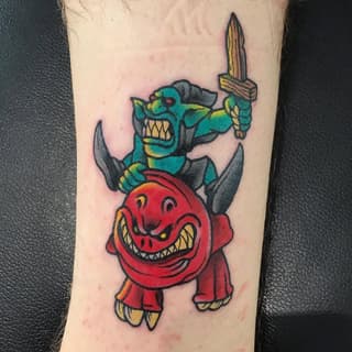 Colorful tattoo on leg featuring a red grinning demon figure with a turquoise character wielding a sword positioned above its head.