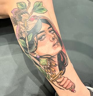 Colorful forearm tattoo featuring a woman's face with closed eyes, a bird perched on flowers, decorative gems, and ornamental elements in pink, green, and gold tones.