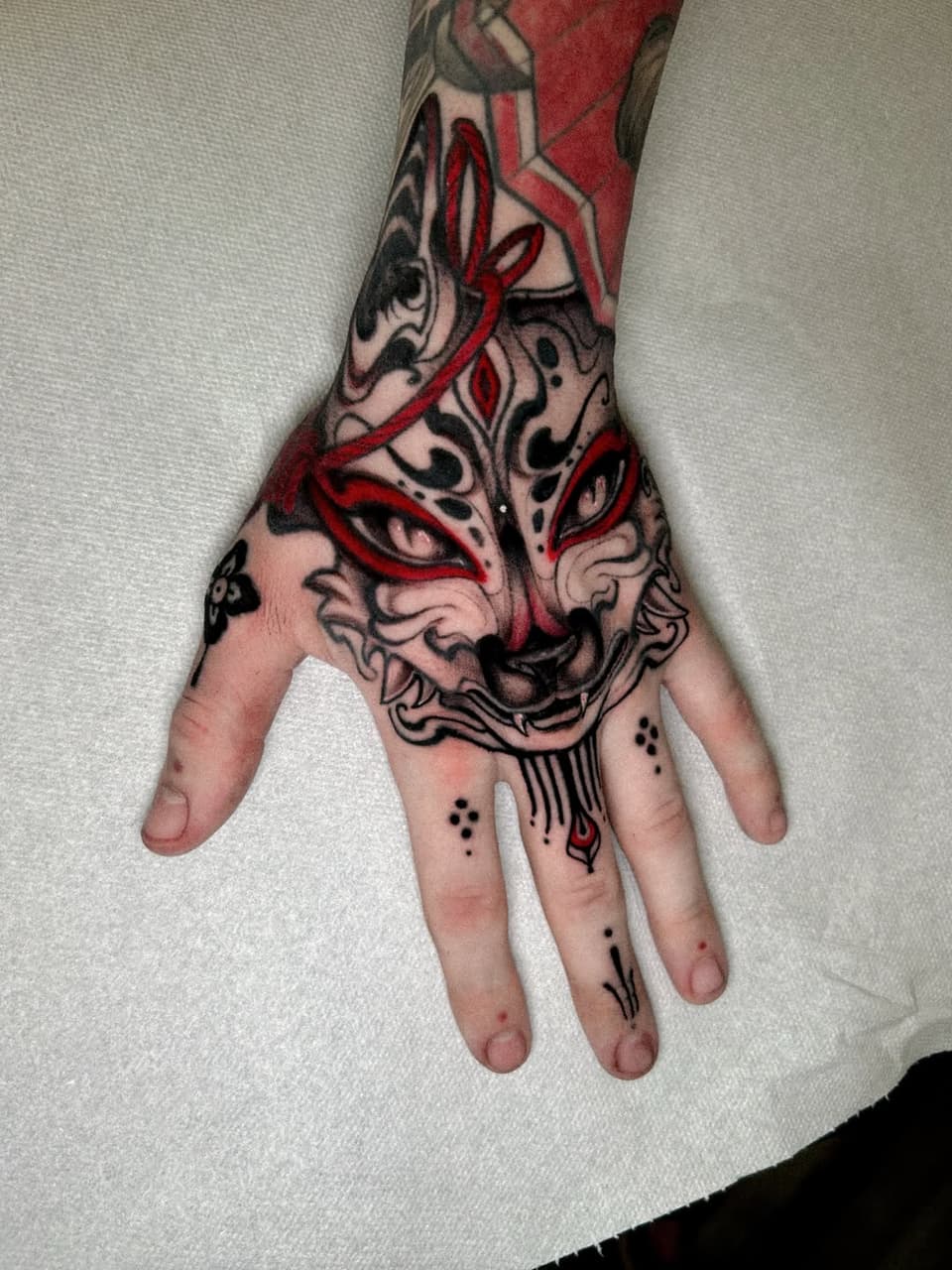 Demon mask tattoo with red and black linework on hand and forearm, featuring ornate facial markings and decorative details on fingers.