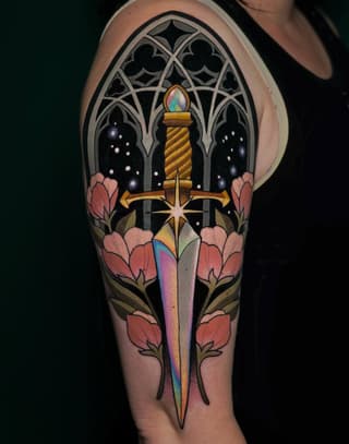 Colorful upper arm tattoo featuring a sword with a rainbow blade, ornate Gothic window, glowing candle, starbursts, pink flowers, and celestial elements on dark skin.