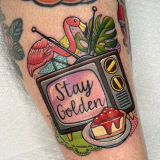 Colorful tattoo on forearm featuring a retro television displaying "Stay Golden" text, with a red bird, monstera plant, strawberry, and decorative elements.