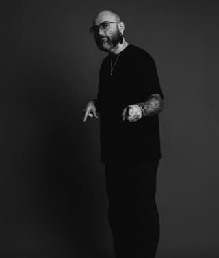 Bald man with full beard wearing glasses and black long-sleeved shirt displays large detailed tattoo covering his entire right forearm and hand against dark background.
