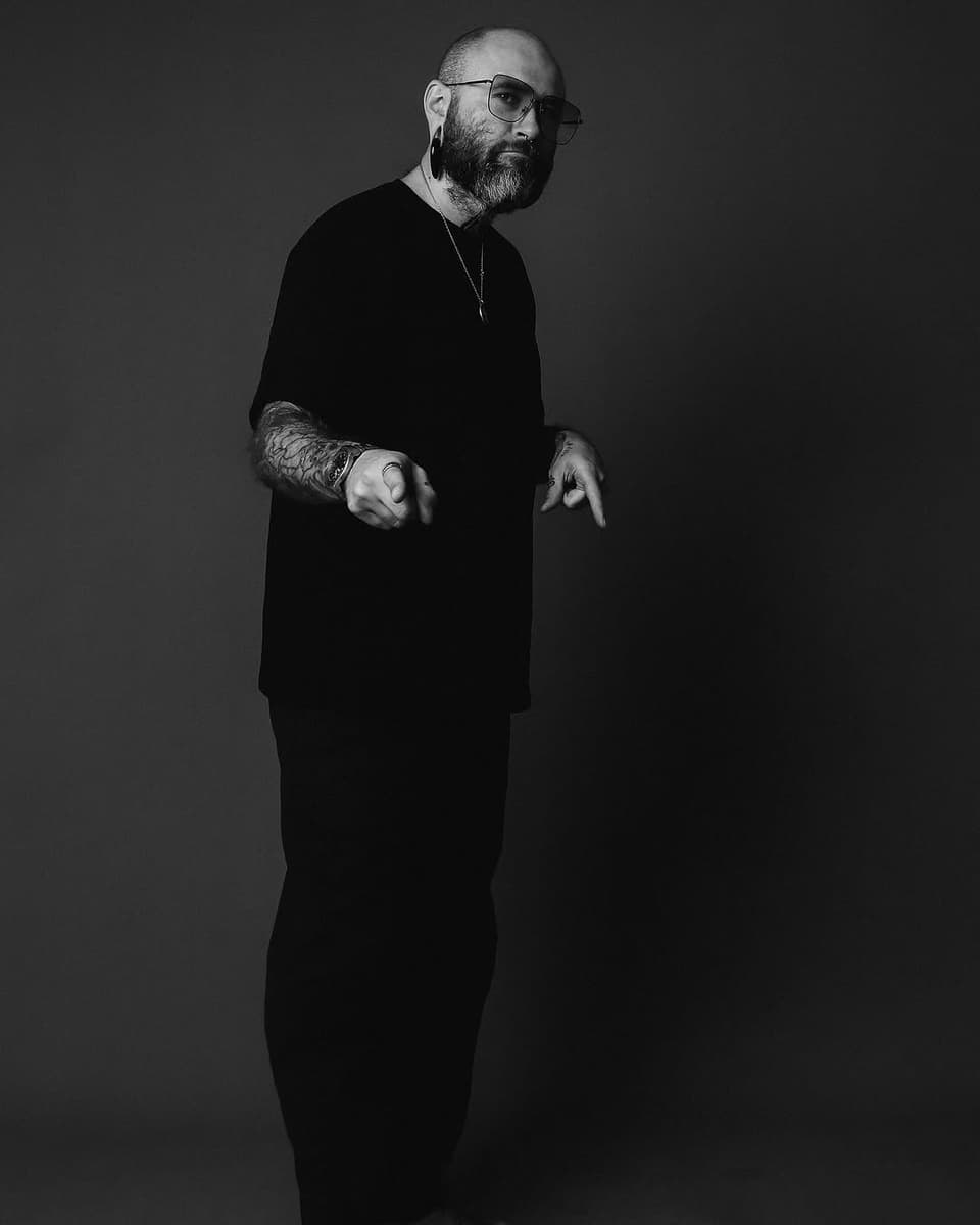 Black and white studio portrait of a bearded man wearing glasses and dark clothing, displaying an arm tattoo on his left sleeve.