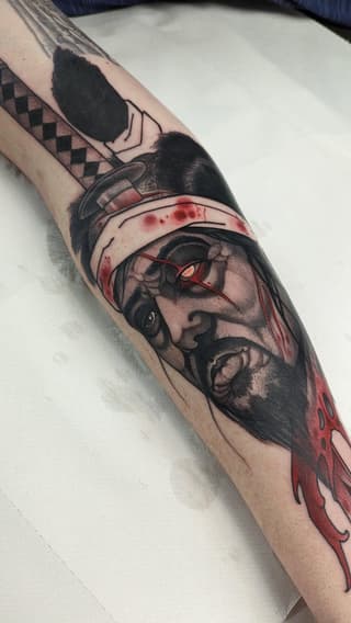 Arm tattoo featuring a bearded man's profile with bandaged head and red eyes, accompanied by geometric patterns and red dripping elements.