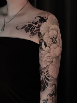 Full arm sleeve tattoo featuring ornate scrollwork and large peony flowers in black and gray ink on pale skin.
