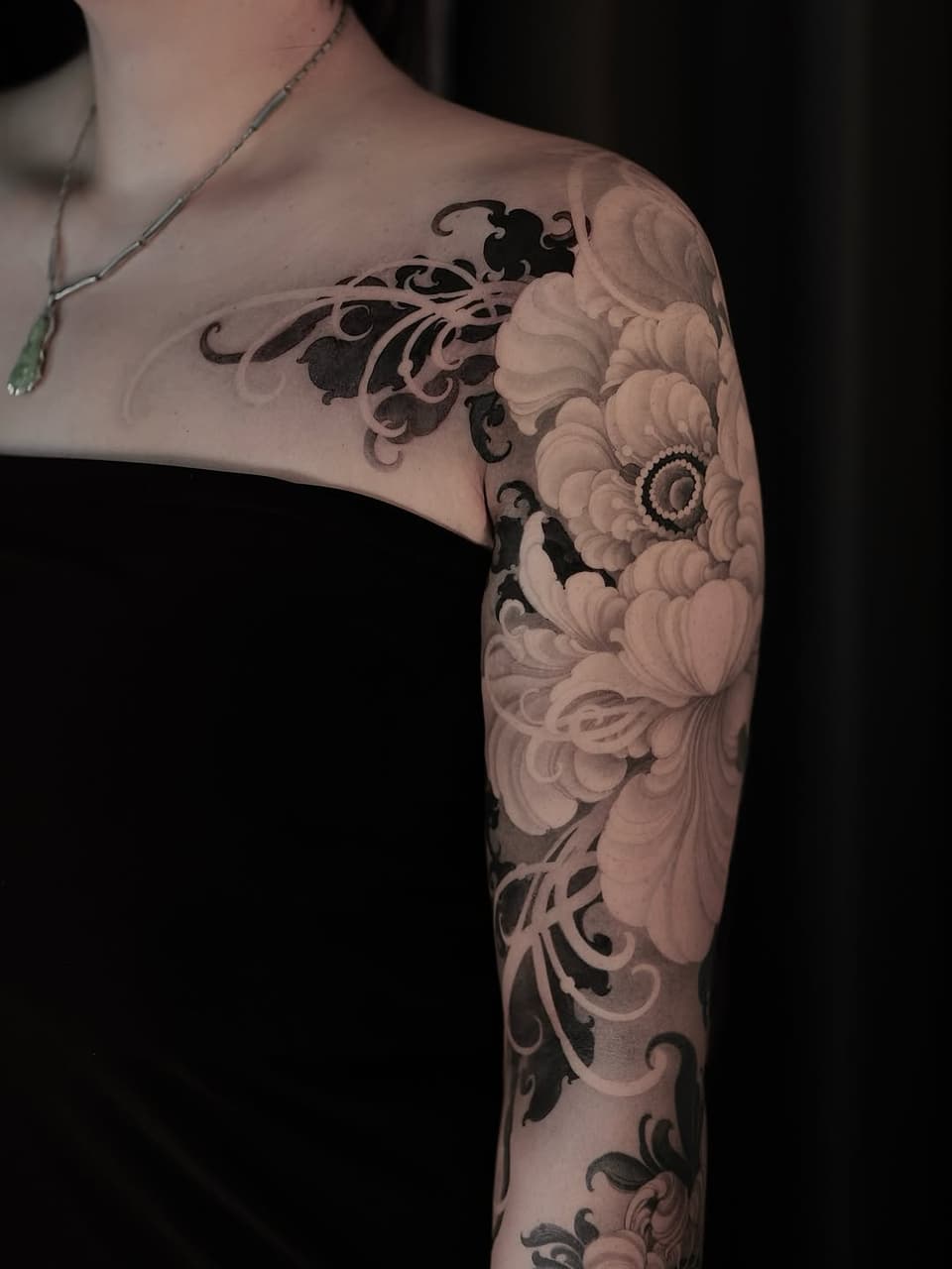 Full arm sleeve tattoo featuring ornate scrollwork and large peony flowers in black and gray ink on pale skin.