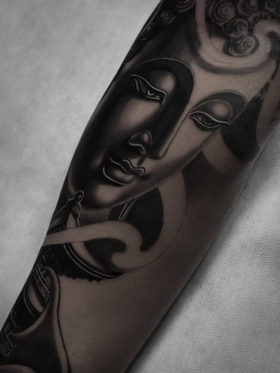 Buddha faces with closed eyes and ornamental headdress details tattooed in black ink on an upper arm, shown against white fabric.