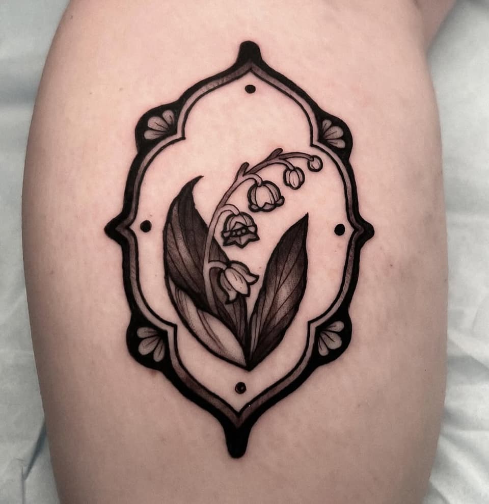 Lily of the valley flowers with leaves in an ornate oval frame with decorative corner elements, tattooed on the thigh in black ink.