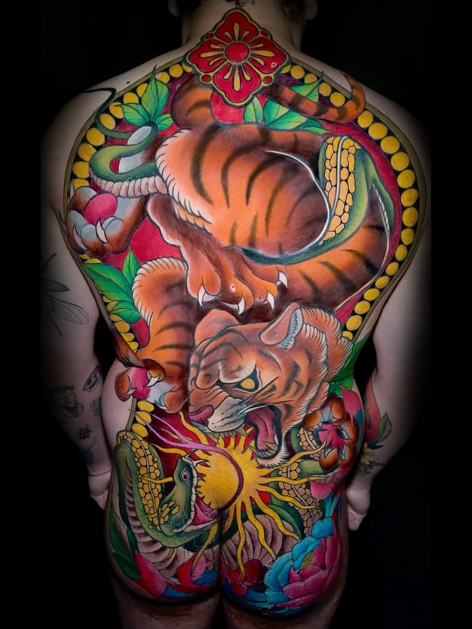 Full back tattoo featuring two tigers with floral and decorative elements in orange, red, yellow, green, and blue colors.