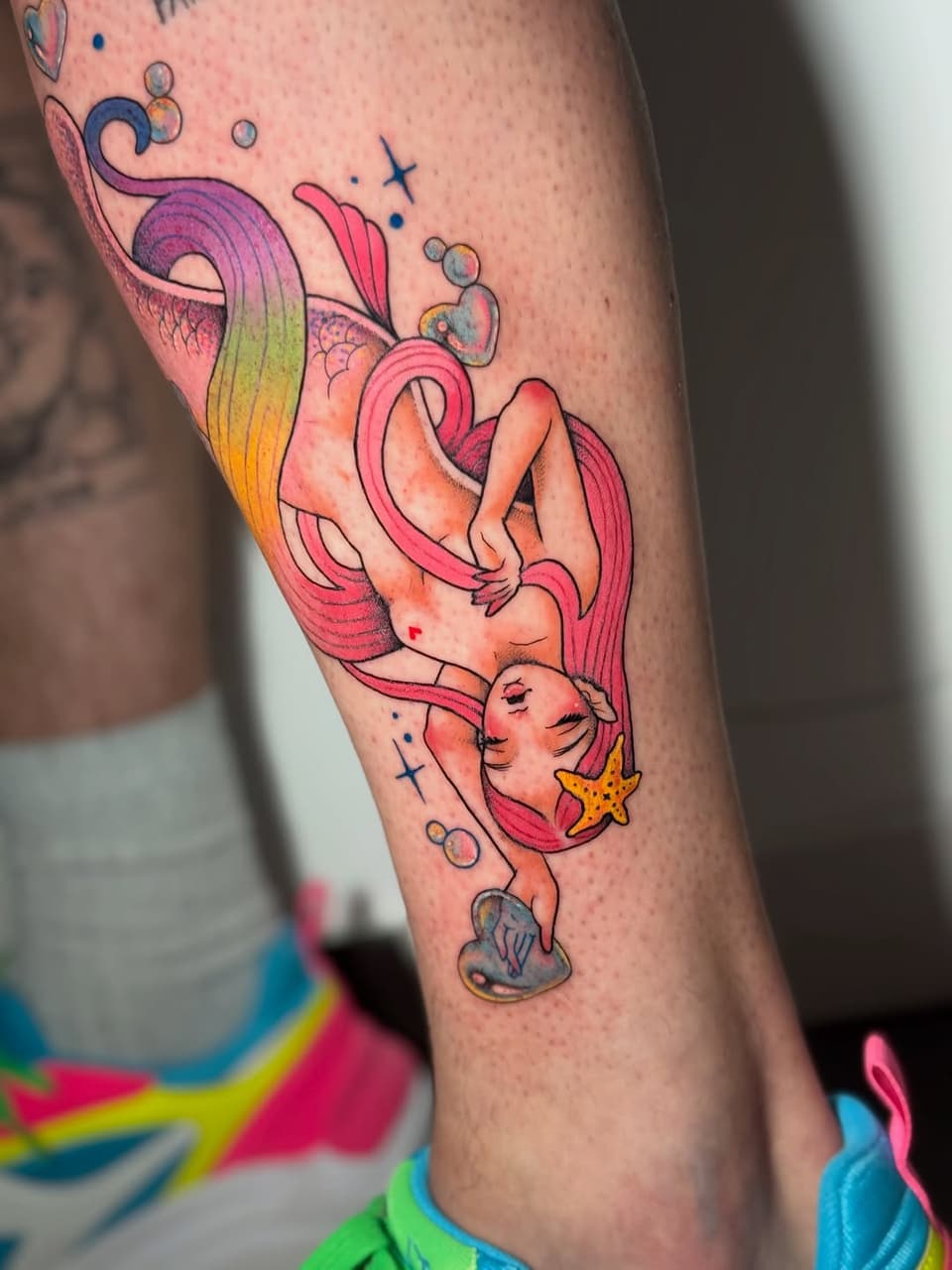 Mermaid with flowing pink and orange hair holding a starfish, surrounded by bubbles and small stars, tattooed on the calf.