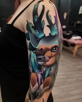 Colorful shoulder tattoo of a deer with teal antlers, brown face, purple lower body, and crystalline geometric elements surrounding it.