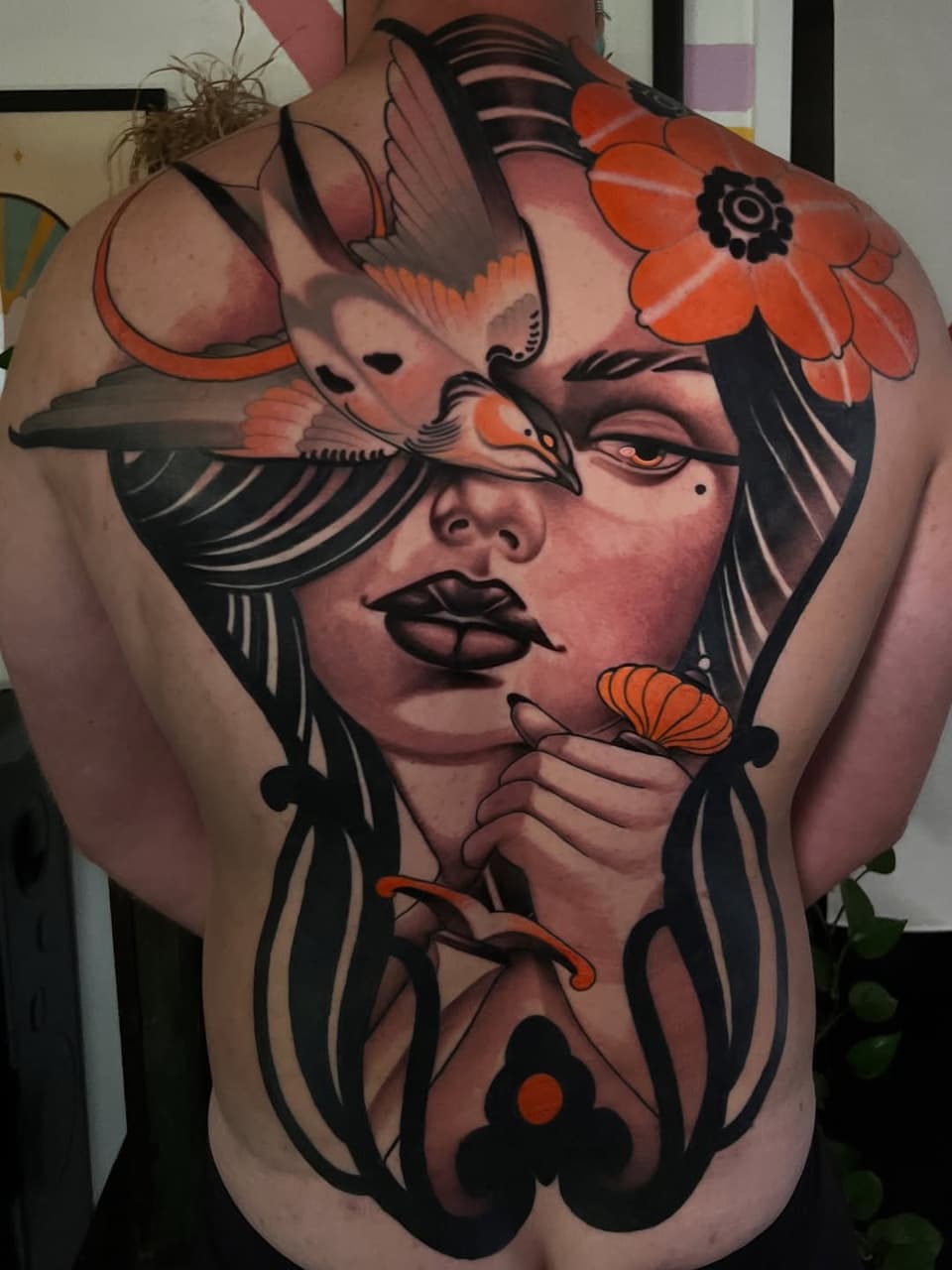 Large-scale back tattoo featuring a woman's face with birds and orange flowers, bold black linework and warm skin-tone shading throughout.