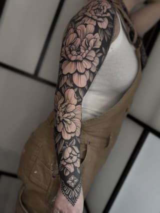 Full-sleeve floral tattoo featuring layered peonies and decorative elements in black ink on a person's arm.