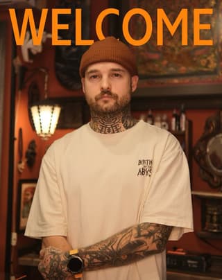 Tattooed man wearing cream t-shirt displays extensive neck and arm tattoos in black ink with geometric and figurative designs.
