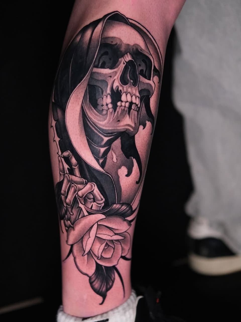 Grim reaper skull with hooded cloak and skeletal hand gesture, accompanied by roses at the base, tattooed on the lower leg in black and pink ink.