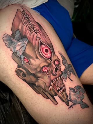 Detailed skull tattoo with fish wings, red eyes, and small birds in flight, positioned on the upper arm in color and black ink.