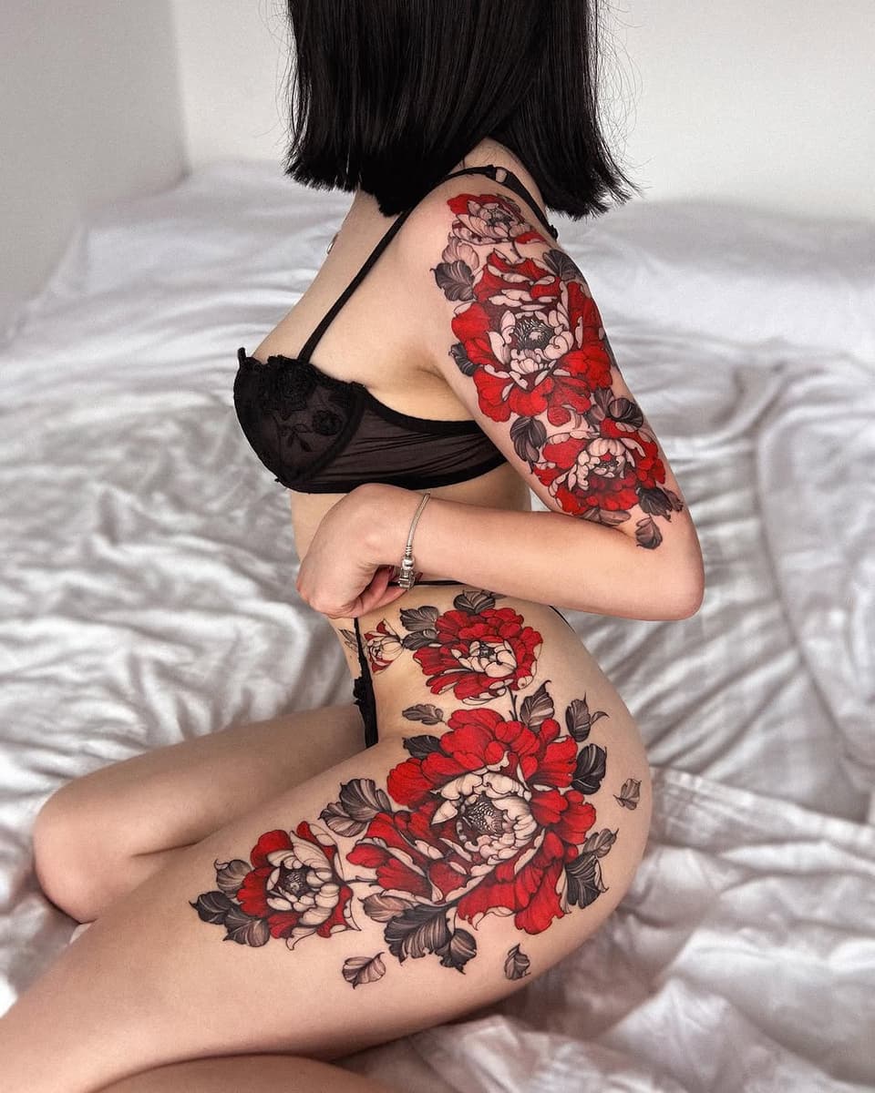 Woman displaying large red and cream floral tattoos with dark foliage covering both thighs and upper arm, positioned on white bedding.