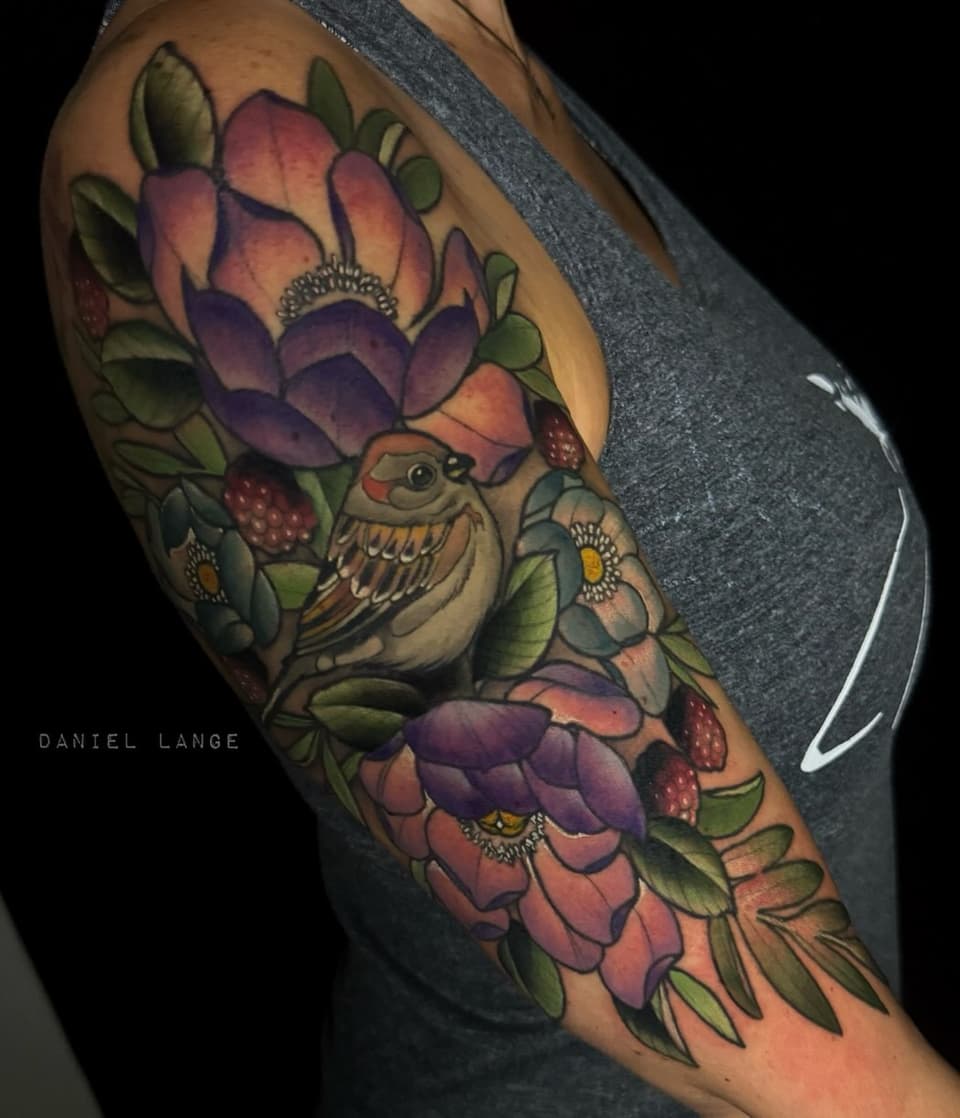 Colorful shoulder tattoo featuring an owl surrounded by purple and pink flowers, green leaves, and red berries on brown skin.
