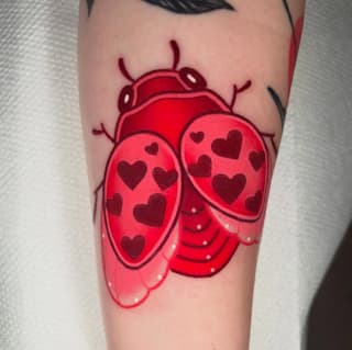 Bright red ladybug tattoo with dark heart patterns on wings, antennae, and segmented body, positioned on upper arm.