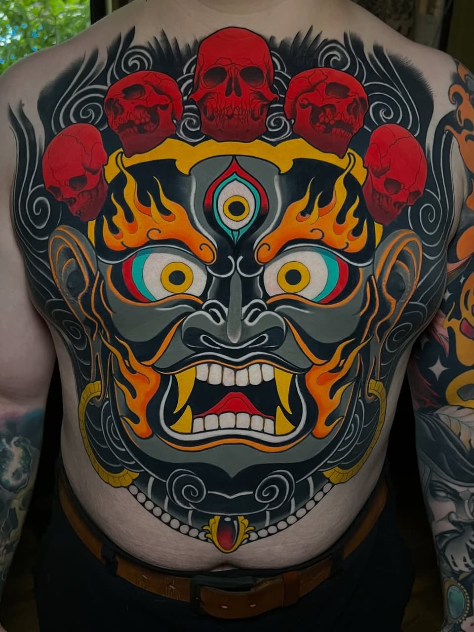 Colorful demon face tattoo covering the back with red skulls above, flame details, large expressive eyes, and ornamental swirls in red, black, orange, yellow, and teal.