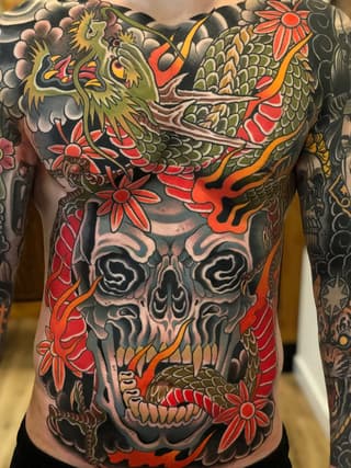 Colorful chest and torso tattoo featuring a skull with dragons, flowers, and ornamental designs in red, green, gold, and black ink.