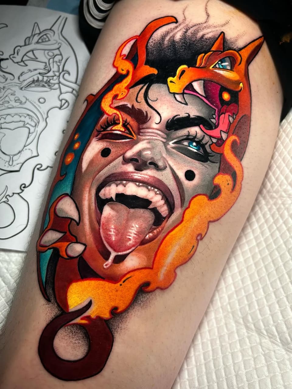 Colorful portrait tattoo on upper arm featuring a face with tongue out, surrounded by red and orange flame designs and decorative scrollwork elements.