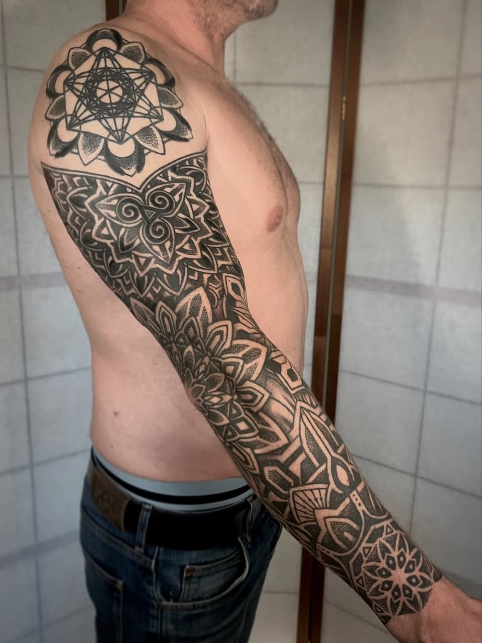 Man displaying a full arm sleeve tattoo featuring geometric mandala and floral patterns in black ink covering shoulder to wrist.