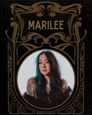 Portrait of a woman with long dark hair and teal streaks displaying colorful floral and geometric tattoos on both shoulders and arms.