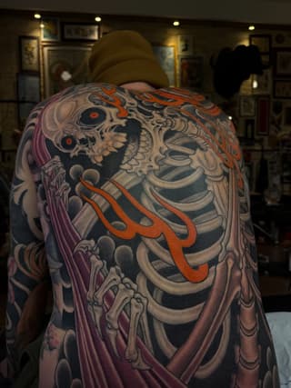 Back and shoulder tattoo featuring a skull with skeletal ribcage, orange flame details, and purple draped fabric elements in multiple colors.