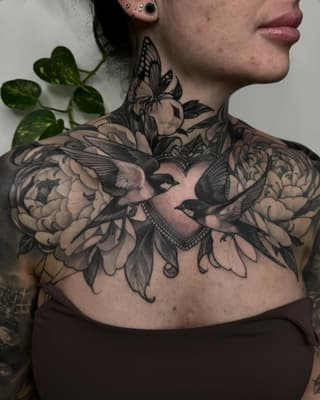Large chest and shoulder tattoo featuring birds, flowers, and ornamental details in black ink on pale skin, displayed on a female model.