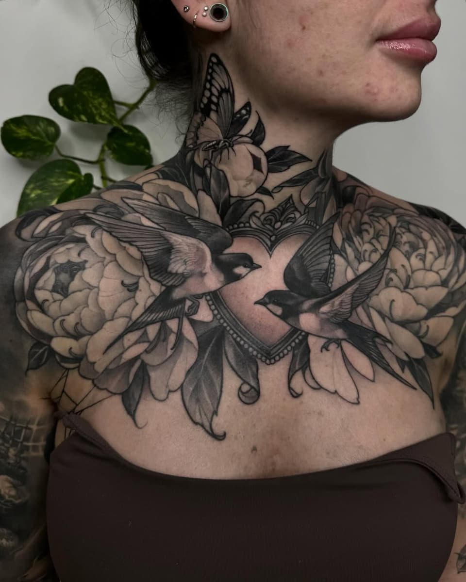 Large chest and shoulder tattoo featuring birds, flowers, and ornamental details in black ink on pale skin, displayed on a female model.