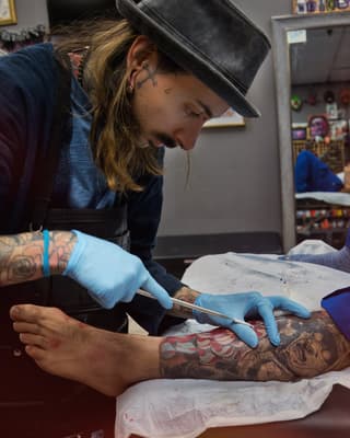 Tattoo artist in black hat and blue gloves applies red and black ink designs to client's forearm during studio session.