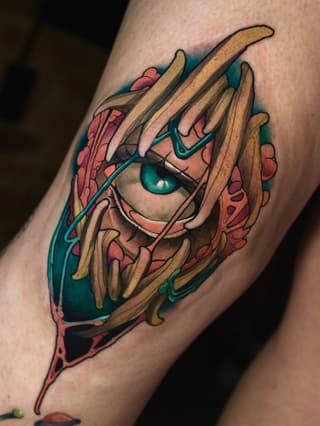 Colorful tattoo on an arm featuring a face with teal eyes and long pointed fingers, surrounded by coral and teal accents.