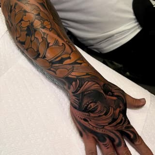 Detailed sleeve and hand tattoo featuring botanical vines, ornamental flowers, and a decorative face design in black and brown ink on brown skin.