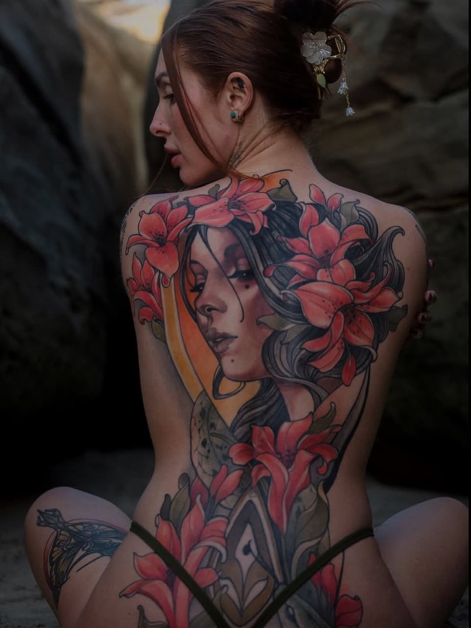 Back tattoo featuring a woman's portrait with flowing hair surrounded by large red flowers and decorative elements in red, orange, and black ink.
