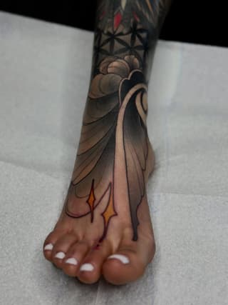 Detailed leg and foot tattoo featuring a draped figure with geometric patterns, shading in black and tan tones with red and yellow accents.