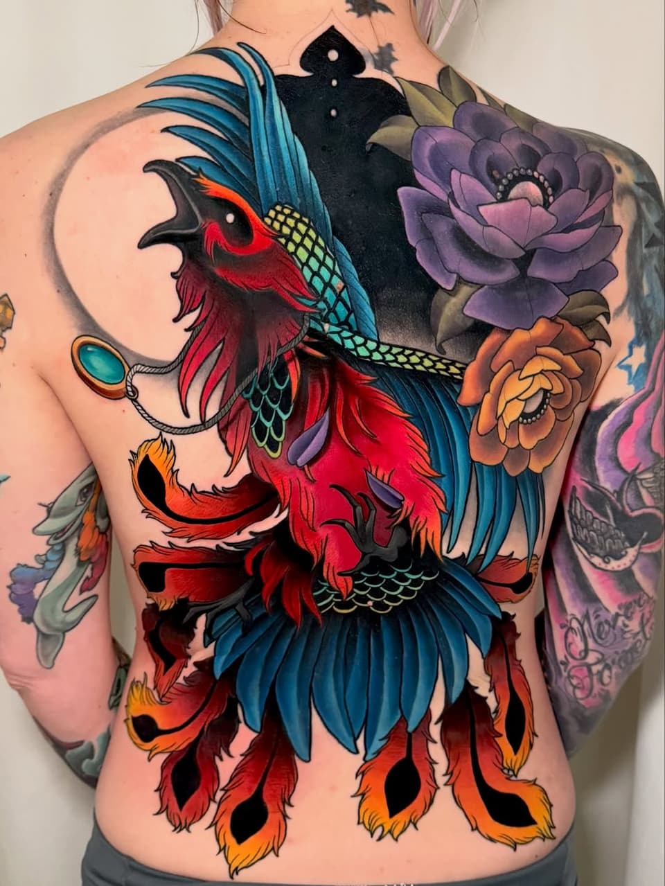 Colorful phoenix tattoo on back with red and blue plumage, turquoise wing details, purple and orange flowers, and a teal gemstone pendant.