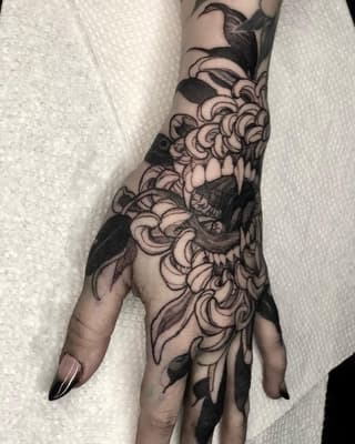 Hand and forearm tattoo featuring ornamental floral and leaf designs with curved scrollwork patterns in black ink on pale skin.