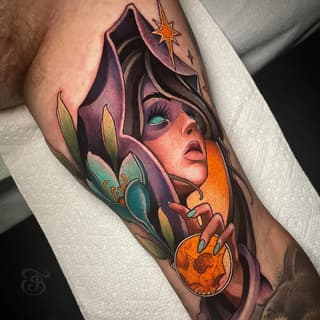 Colorful arm tattoo featuring a hooded figure with turquoise eyes holding an orange orb, surrounded by purple petals and a golden star.