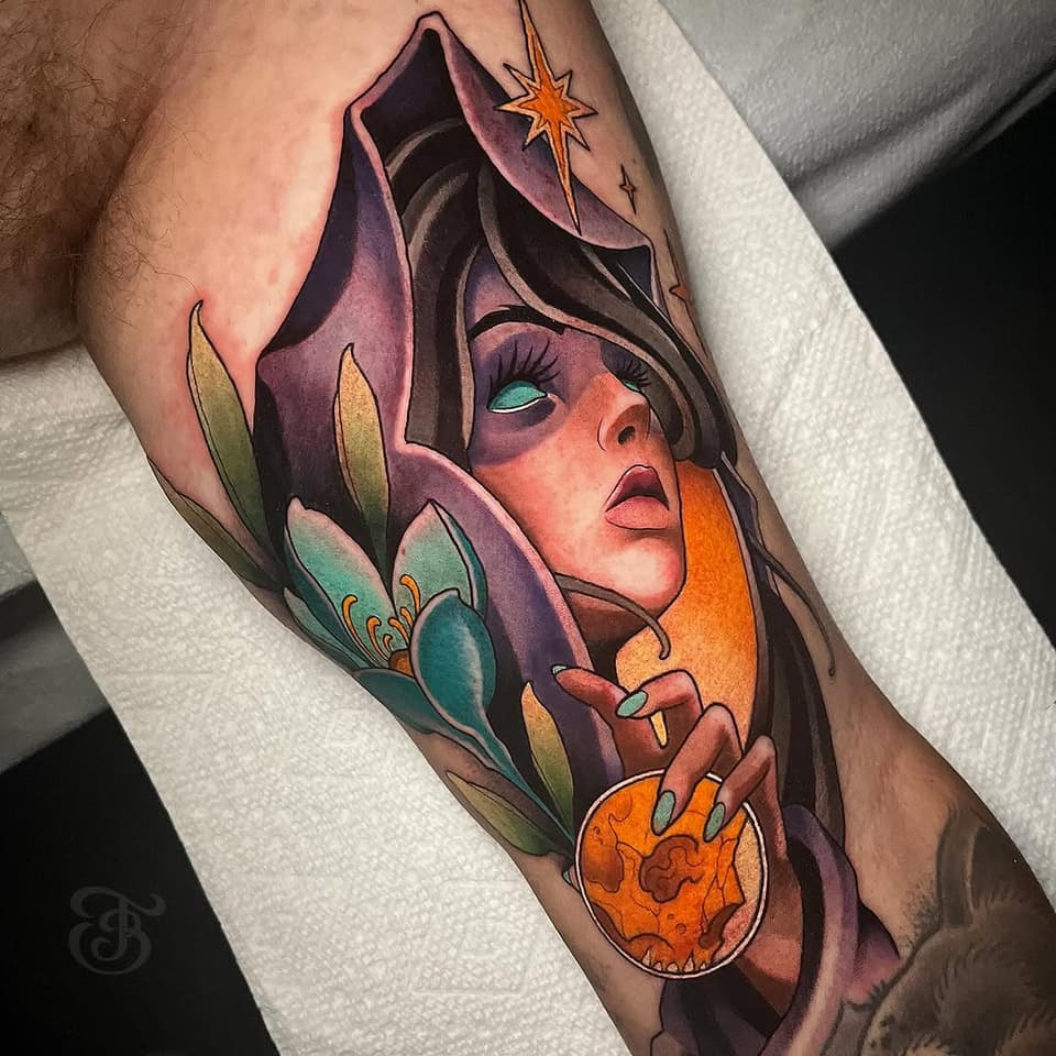 Colorful arm tattoo featuring a hooded figure with turquoise eyes holding an orange orb, surrounded by purple petals and a golden star.